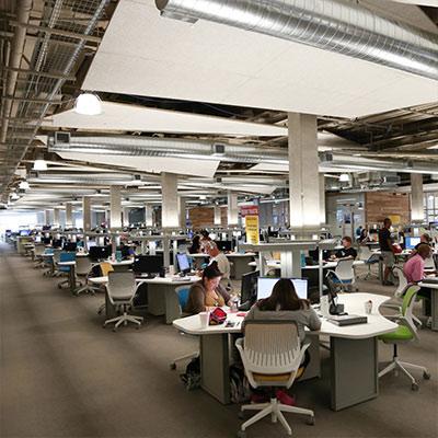 A large, open workspace filled with students sitting at desks with computers, working individually and in small groups. The room has modern lighting and exposed ductwork, giving it a contemporary, industrial feel.